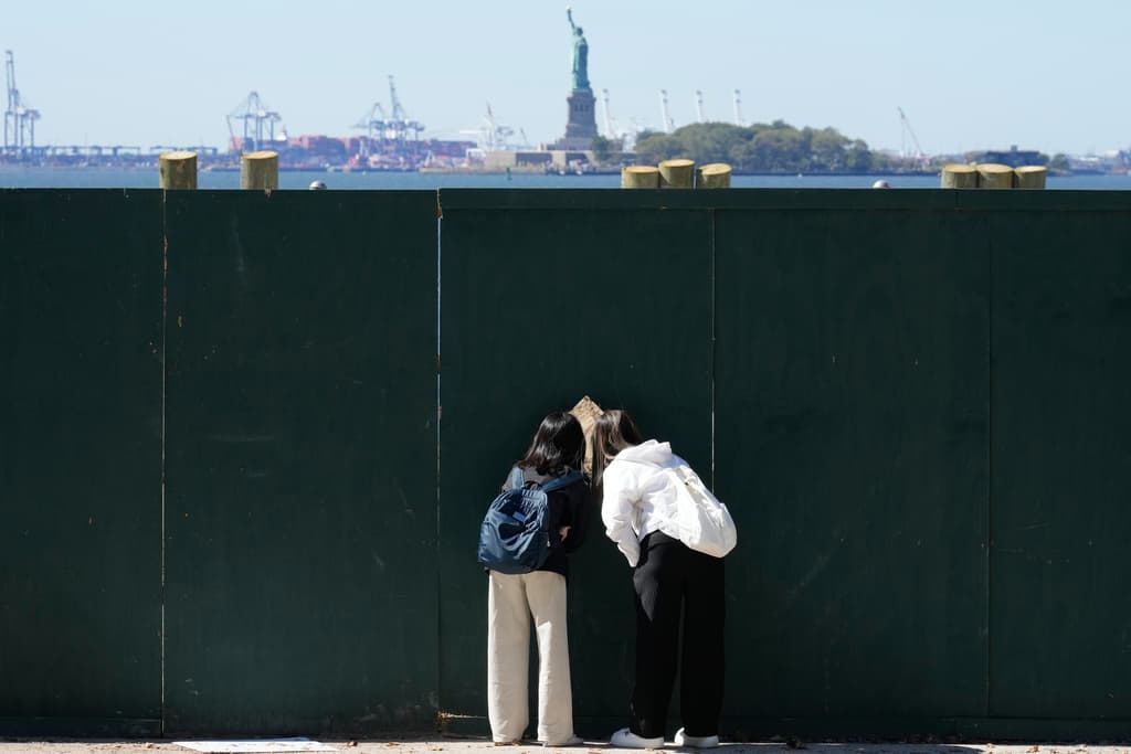 Personas observan a través de una abertura en una cerca para ver la Estatua de la Libertad en Nueva York,
<a href="https://www.univision.com/local/washington-dc-wfdc/cierre-de-gobierno-en-ee-uu-retrasos-en-snap-alza-en-seguros-de-salud-y-un-mes-sin-pagos">durante el primer día del cierre de gobierno</a>, el 1 de octubre de 2025.