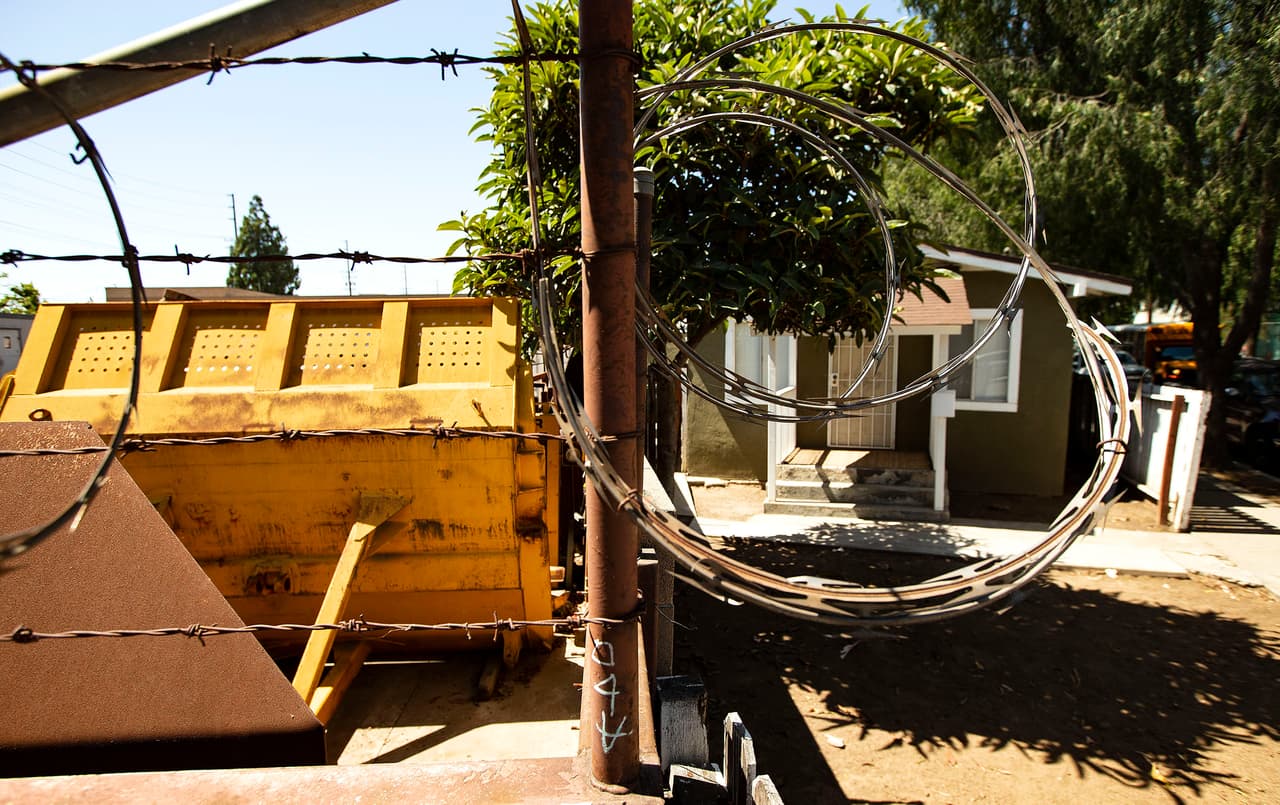 Una reja con alambre de púas cae sobre una pared que separa un negocio industrial y el patio residencial de la casa en la que la familia Garrido vivió en el Barrio Logan, en Santa Ana.