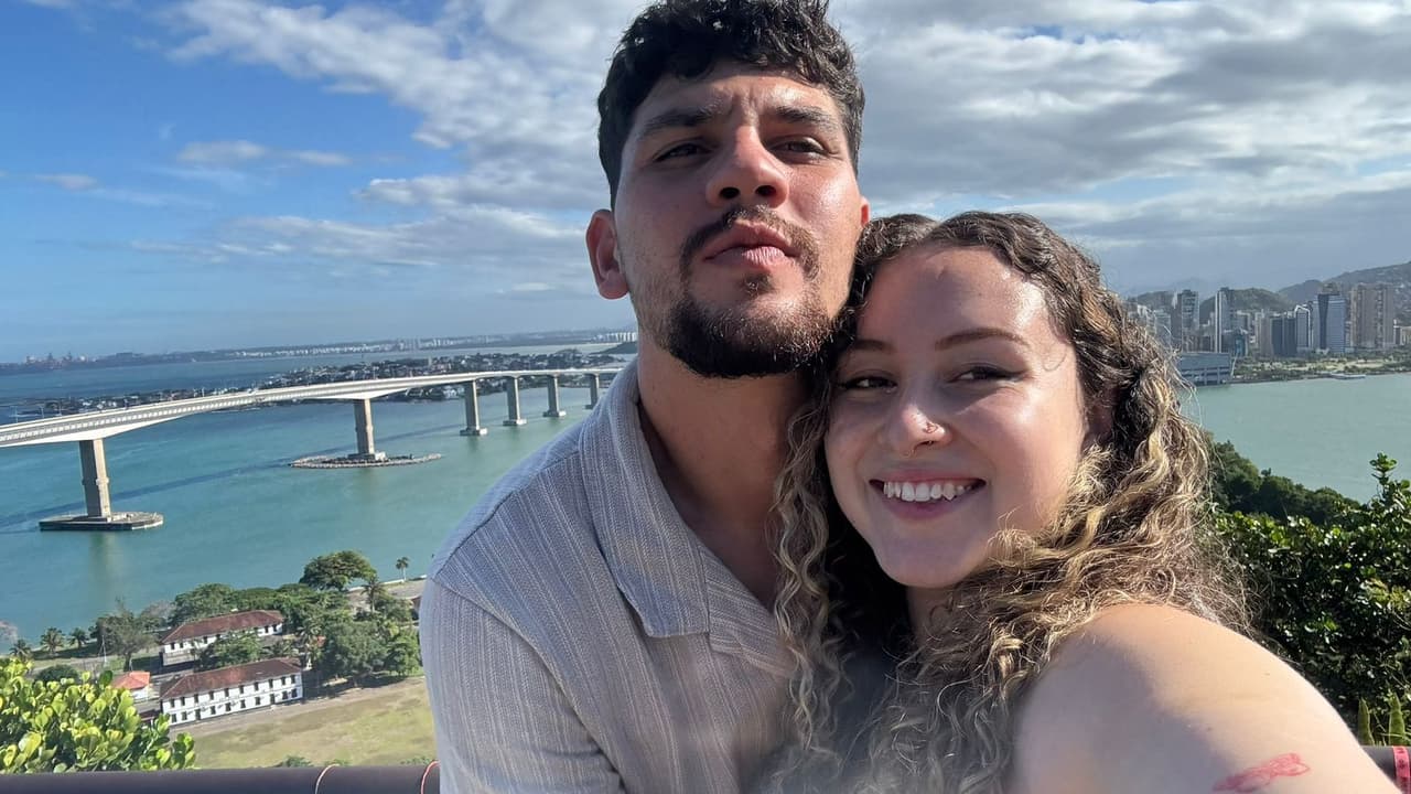Pareja de jóvenes inmigrantes de Colombia y Brasil, Tatiana Melo y Michael Mollulo, posan frente a un puente en Brasil.